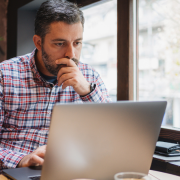 male sitting at cafe table looking at laptop with hand over mouth concerned.