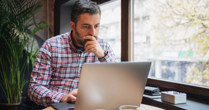 male sitting at cafe table looking at laptop with hand over mouth concerned.